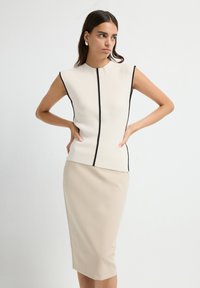 Vrouw in een mouwloze beige ribben top met zwarte biezen en een beige kokerrok, staande met de handen op de heupen tegen een witte achtergrond.