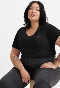 Black V-neck T-shirt made of soft cotton, featuring a small red Levi's logo. Worn with dark gray high-waisted jeans, showcasing arm tattoos.