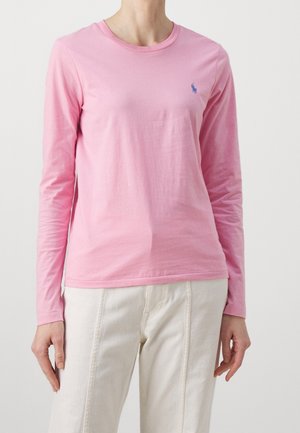 Osoba w jasnoróżowej koszuli z długim rękawem z małym niebieskim wyszywanym logo na klatce piersiowej, w połączeniu z kremowymi spodniami.