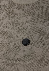 Olivově zelená texturovaná látka s geometrickým vzorem, se černým kulatým knoflíkem s logem, umístěným poblíž výstřihu.