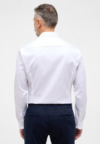Camicia bianca a maniche lunghe in tessuto liscio, con colletto classico e polsini con bottoni, abbinata a pantaloni blu navy.