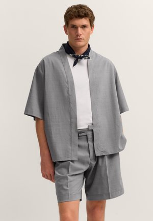Mann trägt locker sitzende graue kurzärmelige Jacke und passende Shorts, weißes T-Shirt und schwarzen Schal um den Hals gebunden, steht vor einfachem Hintergrund.