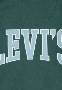 Tessuto verde scuro con testo ricamato di colore blu chiaro che recita "LEVI'S." Le lettere sono in grassetto con contorno bianco, mostrando una texture liscia.