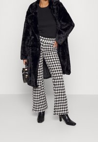 Svart fuskpälsjacka, svart topp, houndstooth-mönstrade byxor i svart och vitt, samt svarta klackstövlar, medan hon håller en liten svart väska.