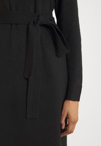 Gros plan d'une robe noire à manches longues avec une ceinture en tissu nouée à la taille et une main reposant sur le côté.