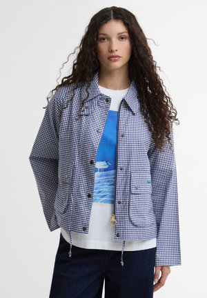 Naine pikkade lokkis juustega, kes kannab sinist-valget ruudulist jopet valge t-särgi kohal, millel on linnu ja vee muster, ning tumedaid pükse.