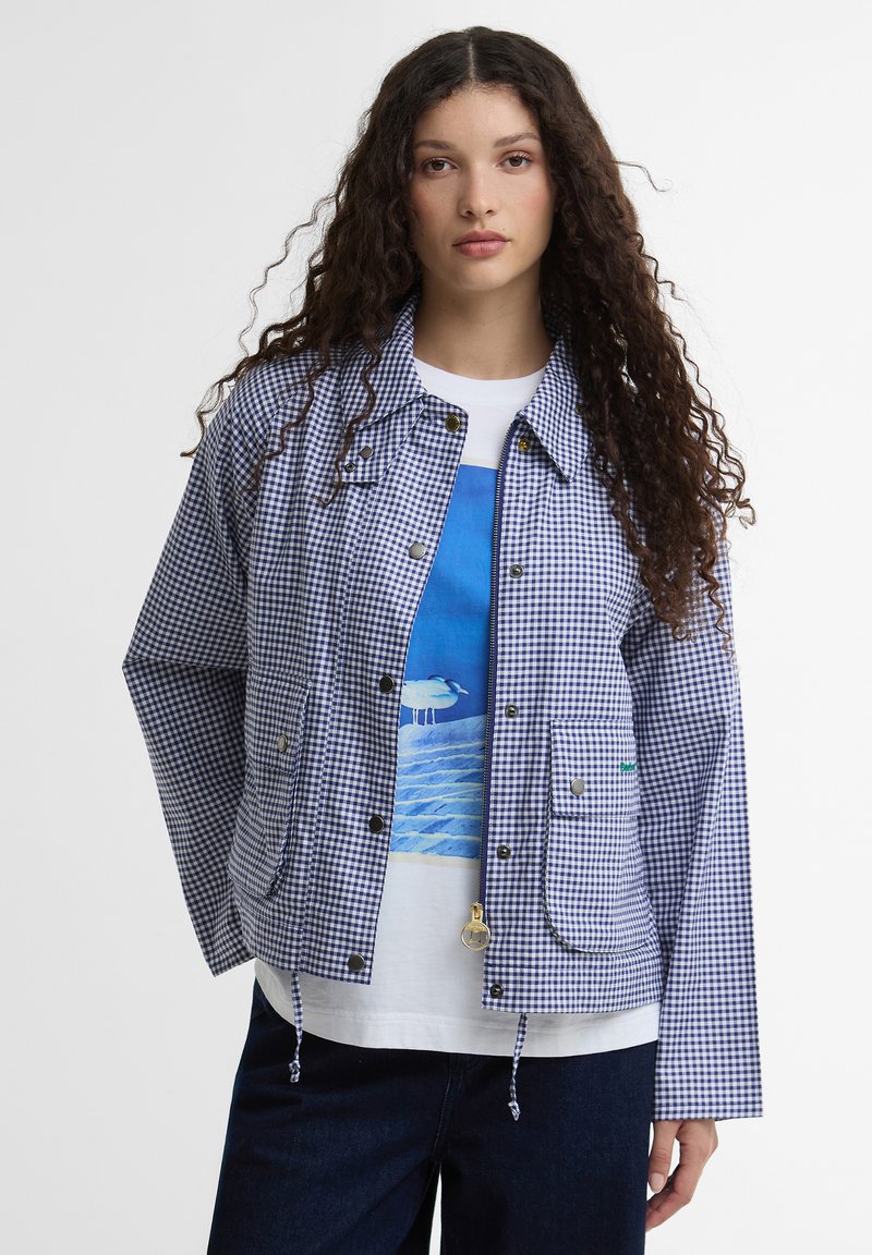 Γυναίκα με μακριά σγουρά μαλλιά, φοράει ένα μπλε και άσπρο καρό σακάκι πάνω από ένα λευκό μπλουζάκι με σχέδιο πουλιού και νερού, και σκούρο παντελόνι.