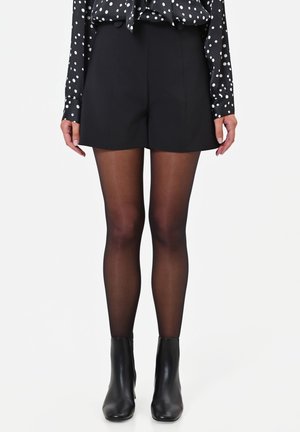 Donna che indossa pantaloncini neri a vita alta, calze nere trasparenti, stivaletti neri e una camicetta nera con pois bianchi.