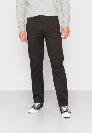 Mann trägt schwarze gerade geschnittene Hose, schwarz-weiße Sneaker und ein hellgraues Langarmshirt, steht auf hellem Boden.