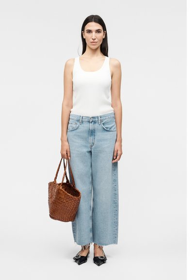 Γυναίκα με ίσια σκούρα μαλλιά, που φοράει λευκό αμάνικο τοπ και ανοιχτό μπλε παντελόνι με φαρδύ πόδι, κρατώντας καφέ πλεκτή τσάντα, μαύρα μυτερά παπούτσια.