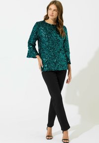 Top in sequins turchese con maniche a tre quarti a campana, abbinato a pantaloni neri aderenti e sandali neri con tacco. Tessuto liscio e lucido per il top.