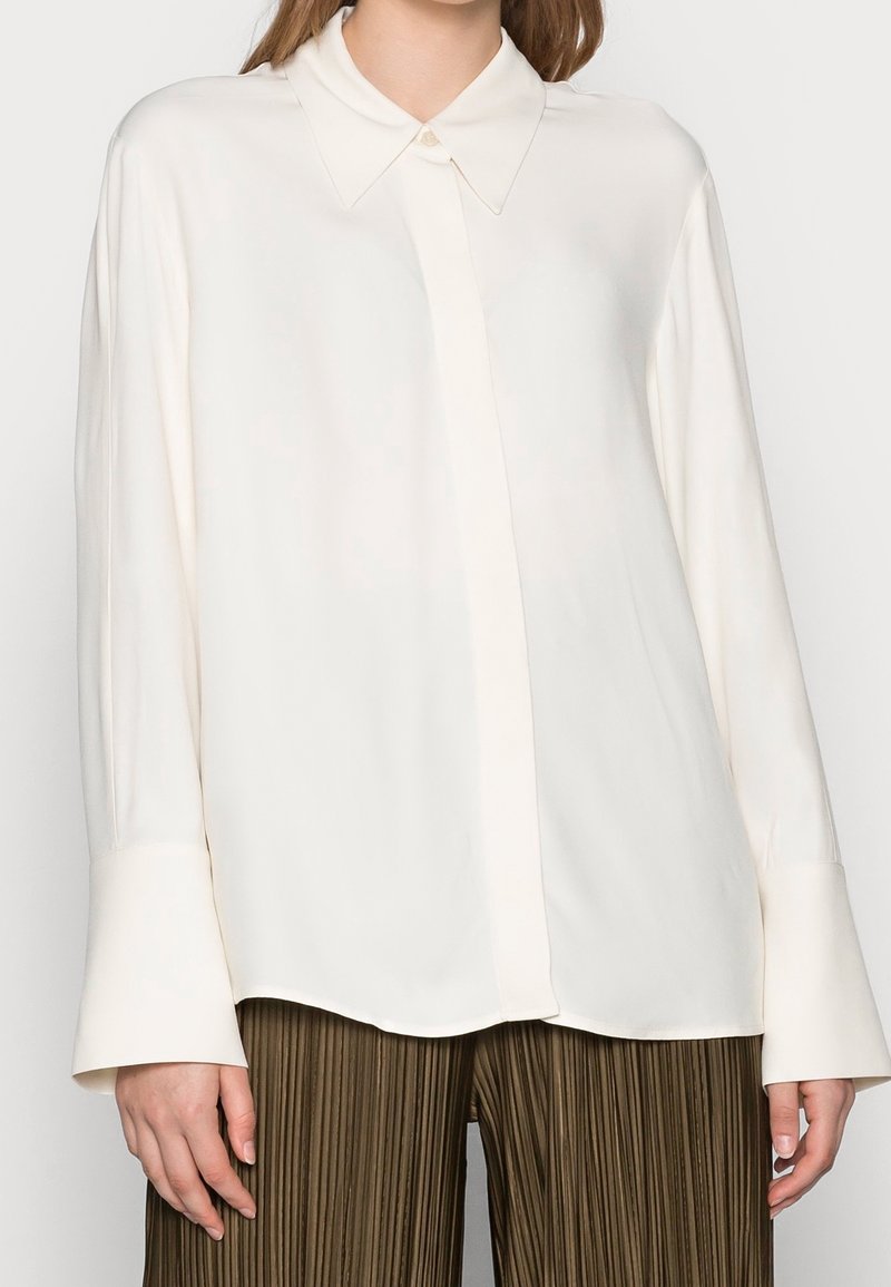 Woman wearing an off-white long-sleeve blouse with wide cuffs and brown pleated pants, cropped at mid-thigh.