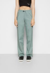 Calças verde claro ajustadas com cintura elástica e fecho com cordão, feitas de um tecido suave, combinadas com sapatos pretos sem fecho.