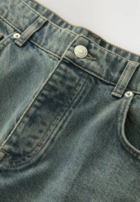 Jean en denim bleu clair, présentant une texture décolorée, des coutures en cuivre, un bouton en métal, et des poches avant avec de subtils détails de couture.