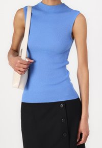 Woman wearing a sleeveless ribbed blue top and black skirt with buttons, carrying a beige shoulder bag on her left shoulder.