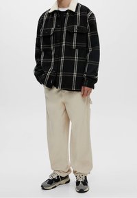 Uomo che indossa una giacca a quadri nera con colletto in pelle bianco, pantaloni beige a gamba larga e sneaker nere e grigie in piedi su uno sfondo bianco.