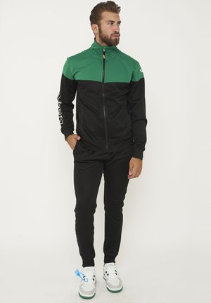 Uomo in piedi con pantaloni neri e giacca verde e nera con zip, indossa scarpe da ginnastica bianche e verdi, guarda di lato con una mano in tasca.