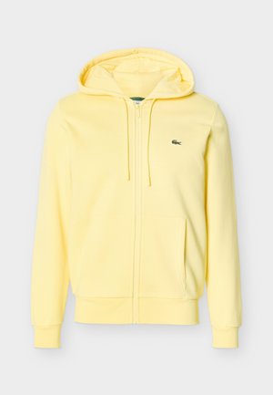 Világos sárga cipzáras kapucnis pulóver húzózsinórokkal, elülső zsebekkel, bordázott mandzsettákkal és egy kis zöld krokodil logóval a mellkason.