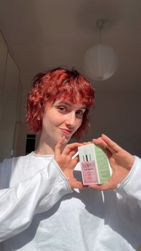 Ung person med rødt hår og hvit langermet skjorte holder Clinique Moisture Surge innpakket hudpleieprodukt i lyst innemiljø.