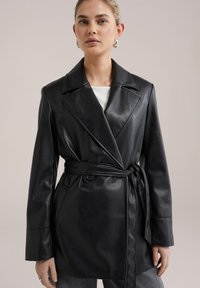 Svart läderjacka med bälte i midjan, stor krage och långa ärmar. Har en mjuk textur och två framfickor.