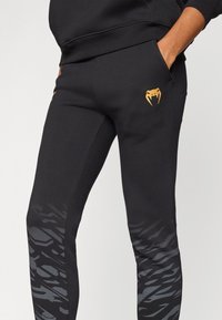 Sorte treningsbukser med slim fit, med et teksturert grått mønster på de nedre bena og en oransje logo på høyre side.