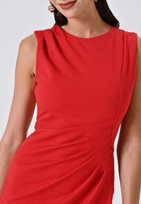 Rotes ärmelloses Kleid mit Rundhalsausschnitt, das mit drapiertem Stoff und dezent gerafften Details an der Seite versehen ist. Weiche Textur.