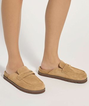 Personne portant des mocassins en daim beige sans lacets avec des semelles marron, debout devant un fond clair uni.