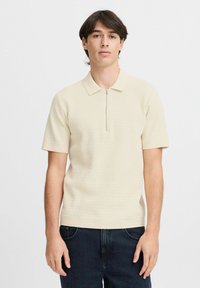 Sweter polo w jasnym beżu z zamkiem przy kołnierzyku, krótkimi rękawami i teksturowanym wzorem. Połączony z ciemnymi dżinsami, minimalistyczny design.
