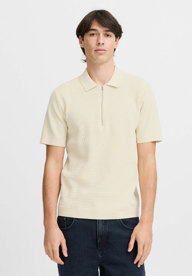 Sweter polo w jasnym beżu z zamkiem przy kołnierzyku, krótkimi rękawami i teksturowanym wzorem. Połączony z ciemnymi dżinsami, minimalistyczny design.