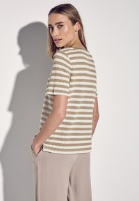 Gestreiftes Strickoberteil mit kurzen Ärmeln in Beige und Creme. Merkmale sind ein runder Ausschnitt und überschnittene Schultern. Kombiniert mit hellbeigen Hosen.