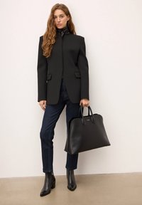 Sort struktureret blazer med høj krave, kombineret med mørke denimjeans. Model holder en stor, glat sort læderhåndtaske. Sorte ankelstøvler.