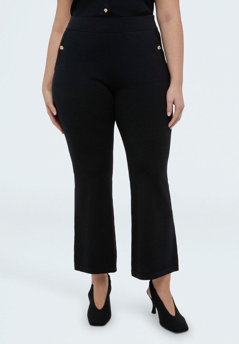 Mujer vistiendo pantalones cortos ajustados negros con detalles de botones dorados y zapatos de tacón negros de punta afilada sobre fondo blanco.