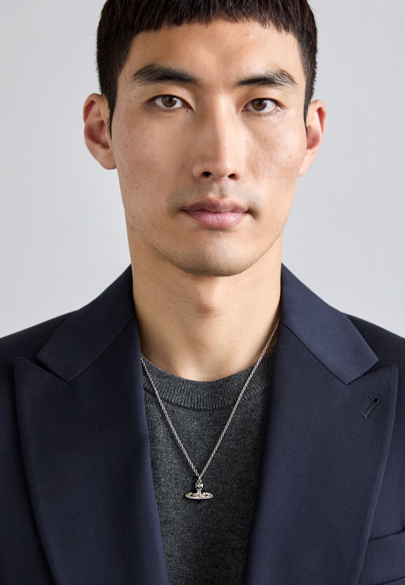 Námořnická sako přes šedý svetr s kulatým výstřihem, zdobené stříbrným náhrdelníkem s jemným přívěskem. Pozadí je neutrálně šedé.