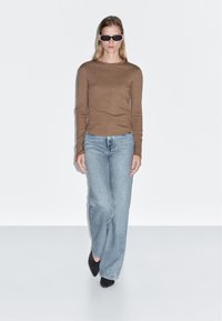 Brauner langärmeliger Strickpullover, hellblaue weit geschnittene Jeans und schwarze spitze High Heels. Das Model trägt schwarze Sonnenbrillen vor einem weißen Hintergrund.