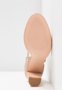 Chaussure à talons haute beige avec un dos ouvert, semelle texturée et coutures subtiles. Forme de talon circulaire, avant plat et sangles croisées qui accentuent le design.