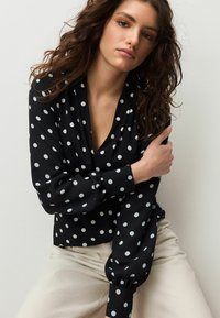 Fekete blúz fehér pöttyökkel, V-nyakkivágással, hosszú, fodros ujjakkal és sima anyaggal. Világos színű nadrággal párosítva.