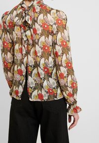 Blumenbluse aus leichtem, gemustertem Stoff mit roten, gelben und pinken Blumen auf schwarzem Hintergrund. Mit Bindeband am Hals und gerafften Bündchen.