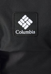 Musta kangas, jossa on suorakulmainen Columbia-logopatchi, jossa on valkoinen geometrinen kuvio. Tekstuuri näyttää sileältä ja kestävältä.