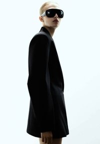 Crni krojeni sako sa struktuiranim dizajnom, uparen s prevelikim crnim naočalama za sunce koje imaju bijeli naglasak na sljepočnicama.