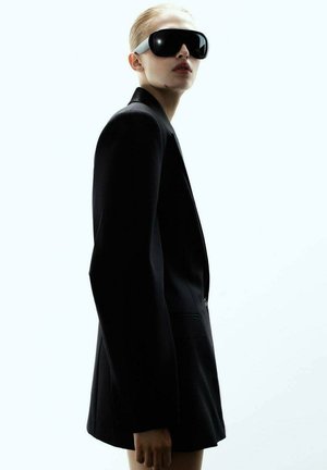 Čierny sakový blejzer so štruktúrovaným dizajnom, kombinovaný s nadrozmernými čiernymi slnečnými okuliarmi so bielym akcentom na rámiku.
