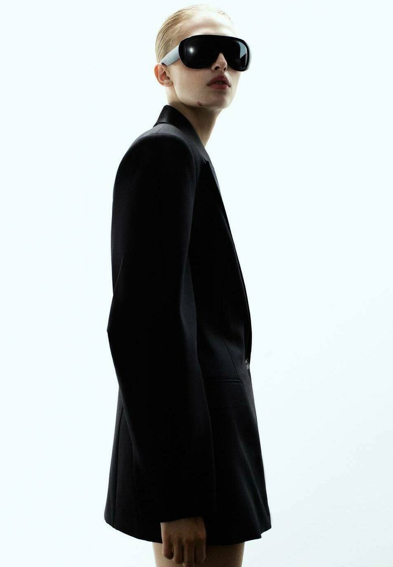 Crni krojeni sako sa struktuiranim dizajnom, uparen s prevelikim crnim naočalama za sunce koje imaju bijeli naglasak na sljepočnicama.