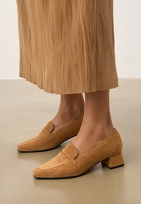Mocassins à talons carrés en daim tan, avec des détails cousus et une petite bande décorative. Les chaussures présentent un bout pointu et une surface texturée.