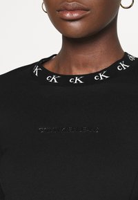 Svart crewneck-topp med en texturerad krage med upprepande "ck"-mönster, präglad "CALVIN KLEIN JEANS"-logotyp på framsidan.