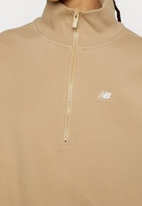 Sweat-shirt beige à fermeture quart de zip avec col côtelé et logo blanc New Balance sur le côté gauche de la poitrine.