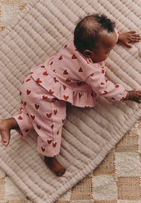 Rosa barnkläder med röda hjärtmönster, bestående av en top med volang och matchande byxor, placerade på en texturerad beige matta.