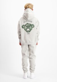Grijze hoodie met een grote groene grafische print van een gestileerd gezicht. Gecombineerd met bijpassende joggingbroek en witte sneakers. Glad materiaal, casual pasvorm.