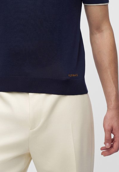 Gros plan d'une personne portant une chemise bleu marine avec « Versace » brodé près de l'ourlet et un pantalon couleur crème, bras droit visible.