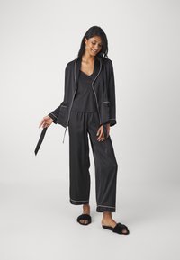 Svart satin pyjamas-set med en långärmad jacka, raka byxor och en v-ringad tanktop. Har tunna vita detaljer och mjuka svarta tofflor.
