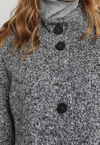 Šedý texturovaný kabát s vysokým límcem, který má tři černé knoflíky. Tkanina má tečkovaný vzor a působí teplým, hustým dojmem.
