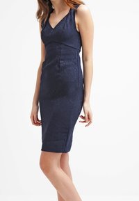 Navy blauwe aansluitende jurk van een gestructureerde stof, met V-hals en mouwloos ontwerp. Kenmerkt zijzakken en heeft een lengte tot de knie.